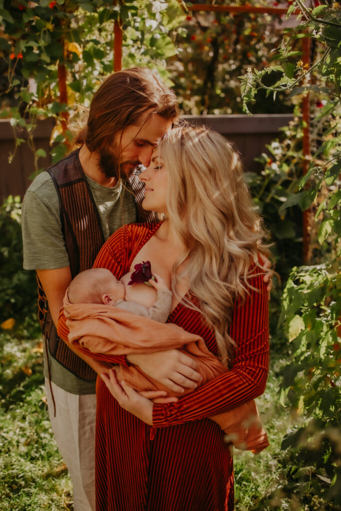 A tender moment of a mother and father embracing their newborn baby in a lush garden setting, the mother cradling the infant closely while the father leans in gently. The image captures the intimacy of early parenthood, highlighting the importance of nurturing moments. Newborn photo shoot Edmonton.