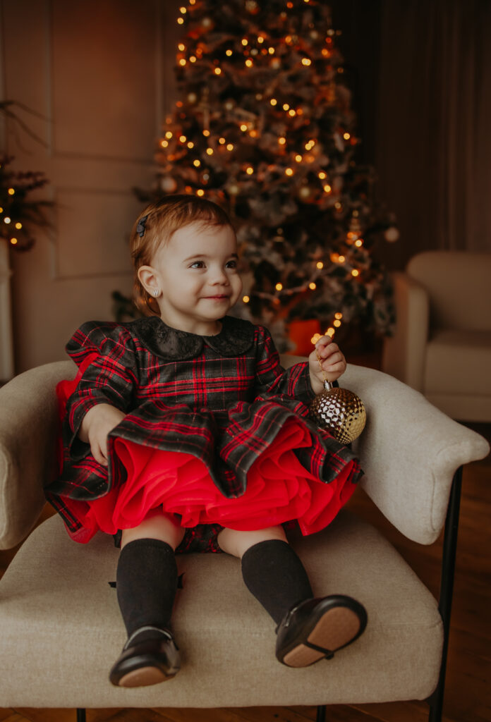 A young child sitting on a beige chair in front of a beautifully lit Christmas tree, wearing a festive red and black plaid dress with a red underskirt, holding a gold ornament, and smiling warmly during a christmas family photo shoot in Edmonton 