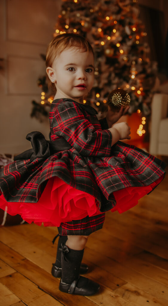 A toddler girl standing in front of a christmas tree in a festive red and black check dress holding a gold christmas tree bulb during a christmas family photo shoot in Edmonton 