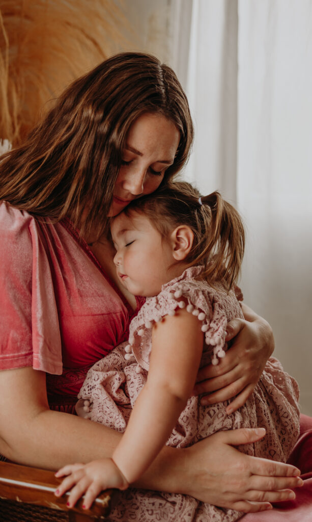 a mom in a pink dress holds her toddler daughter sitting in a chair during her indoor family photo session in Edmonton, Alberta