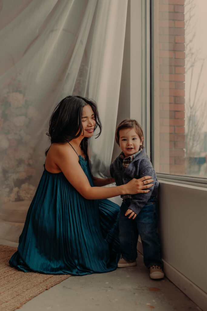 Mom in a blue dress kneels next to toddler son during an indoor Edmonton maternity photo shoot