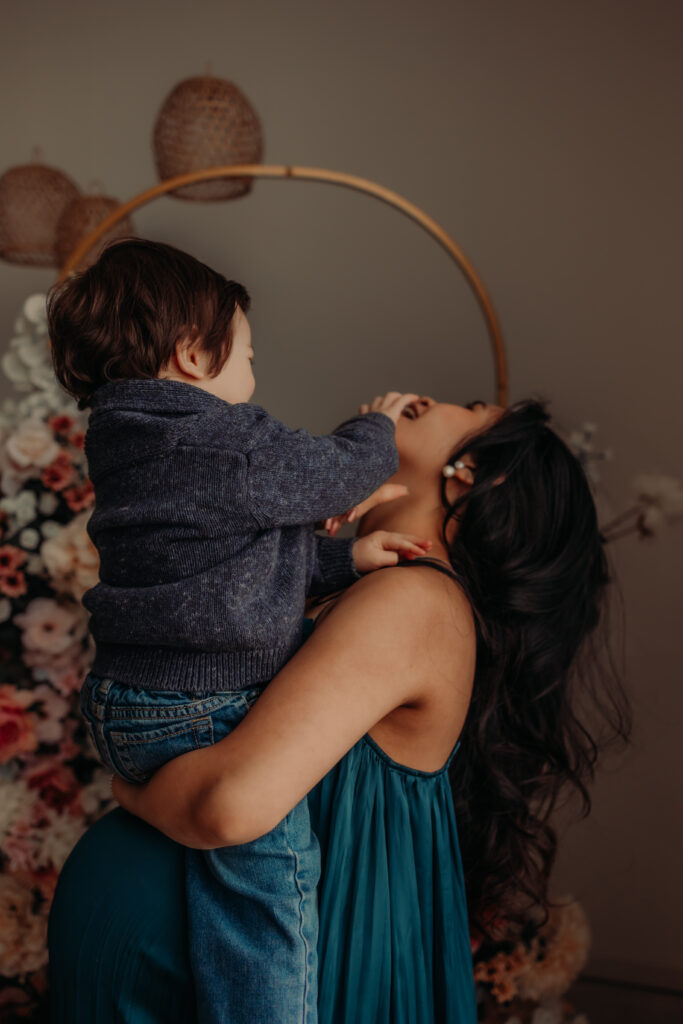 Mom in a blue dress holds toddler son while laughing during Edmonton maternity photo shoot