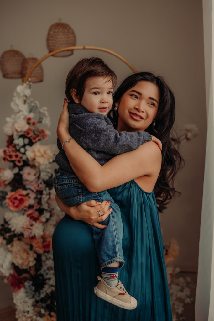 Mom in a blue dress holds toddler son during an indoor Edmonton maternity photo shoot