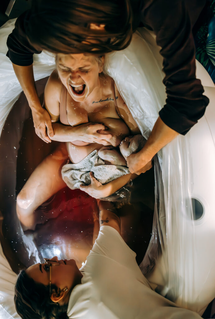 A mother roars with power as she delivers her placenta in a birth tub, surrounded by support. Her partner holds her, while a birth worker reaches in to assist. The raw intensity of labor are captured in this powerful moment.