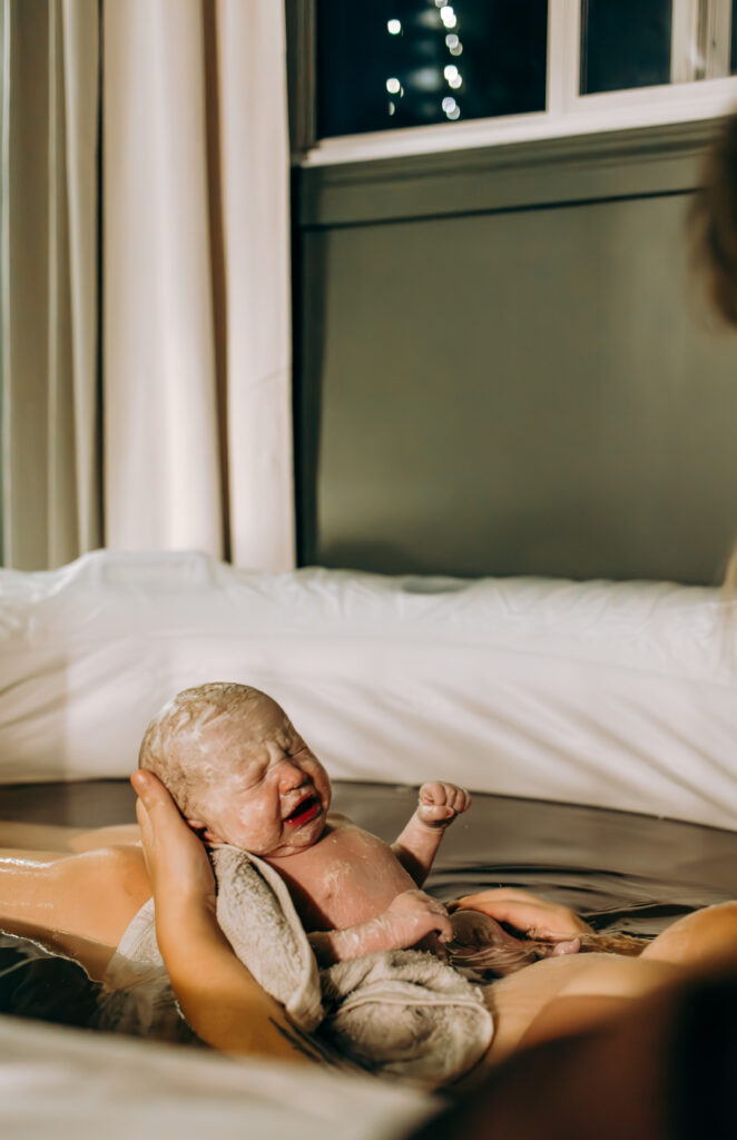 A mother cradles her newborn baby on her chest in a birth tub, surrounded by loving hands. The baby, covered in vernix, is gently touched by both parents, capturing the raw emotion and intimacy of birth. The mother gazes at her baby with love, while her partner leans in close, sharing in the powerful moment.