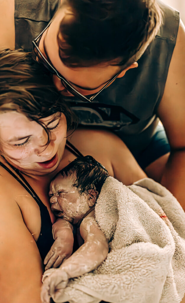 A newborn baby covered in vernix rests on their mother’s chest in a warm birth tub while dad  kisses mom's head. The intimate moment captures the raw beauty of a water birth.