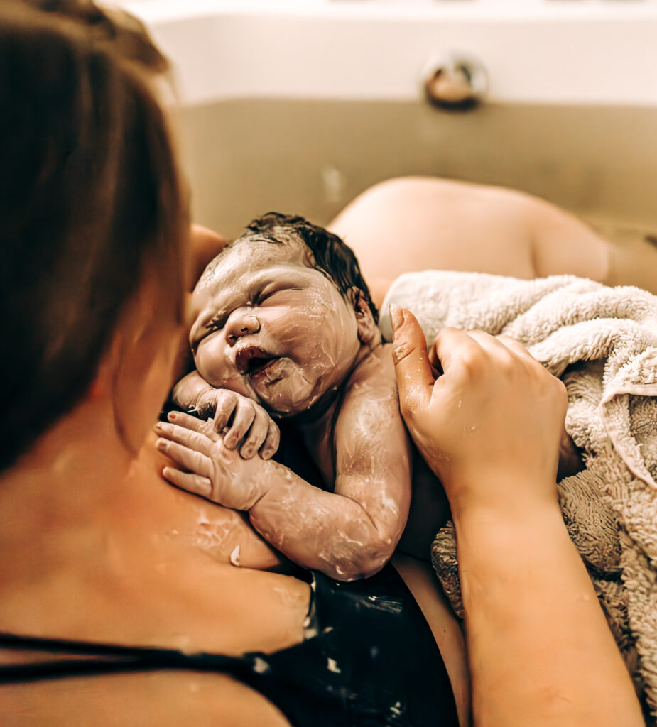 A newborn baby covered in vernix rests on their mother’s chest in a warm birth tub. The baby’s tiny hands clutch the mother’s skin, and the mother’s arm supports them, wrapped in a soft towel. The intimate moment captures the raw beauty of a water birth.