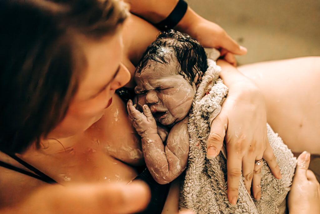 A mother cradles her newborn baby on her chest in a birth tub, surrounded by loving hands. The baby, covered in vernix, is gently touched by both parents, capturing the raw emotion and intimacy of birth. The mother gazes at her baby with love, while her partner leans in close, sharing in the powerful moment.