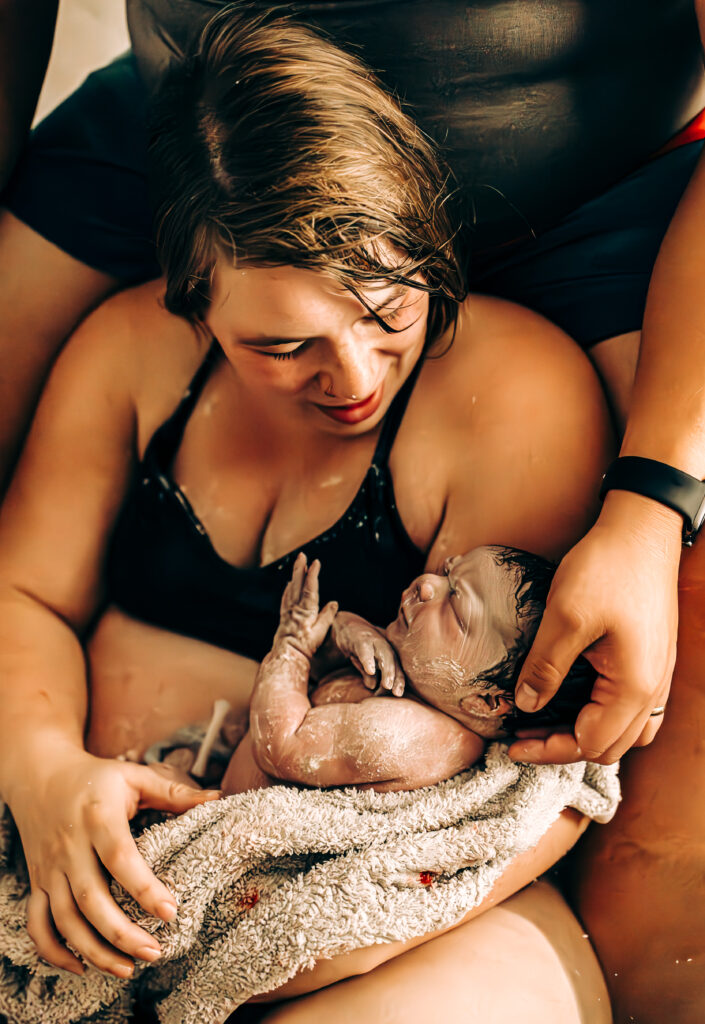 A newborn baby covered in vernix rests on their mother’s chest in a warm birth tub. The baby’s tiny hands clutch the mother’s skin, and the mother’s arm supports them, wrapped in a soft towel. The intimate moment captures the raw beauty of a water birth.