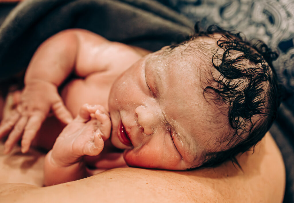 a baby still covered in vernix rests on his mother's chest after birth wrapped in a towel