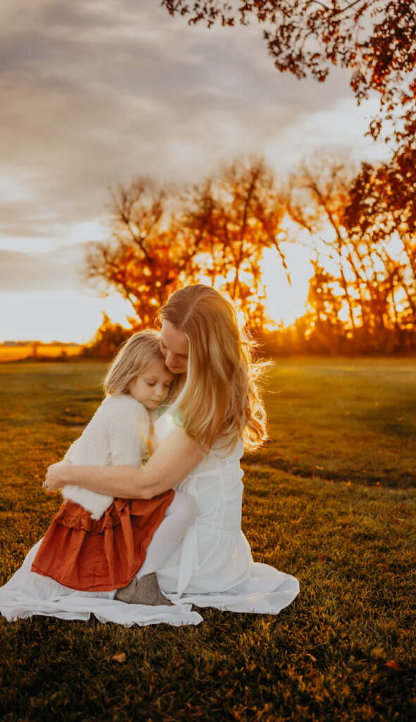 a mother holds her daughter tenderly on her lap against the setting sun during an fall edmonton family photo session