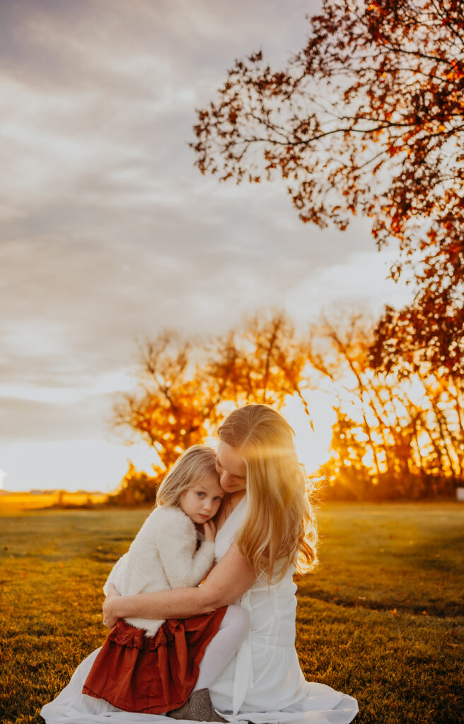 a mother holds her daughter tenderly on her lap against the setting sun during an fall edmonton family photo session