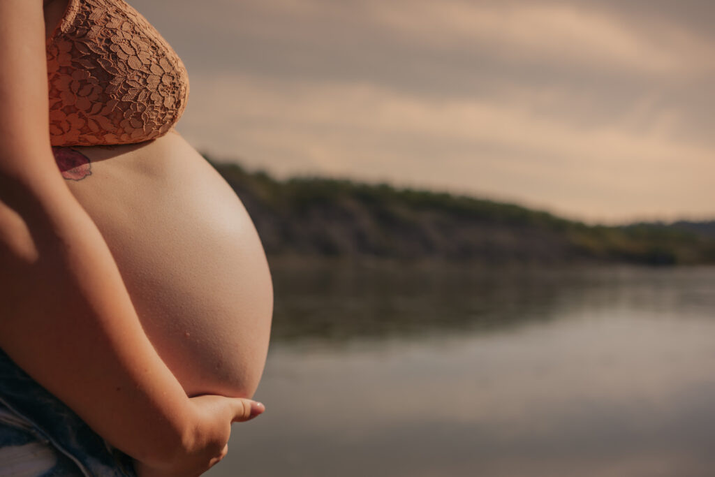 pregnant woman poses in jeans and bra by river during outdoor maternity photos in Edmonton