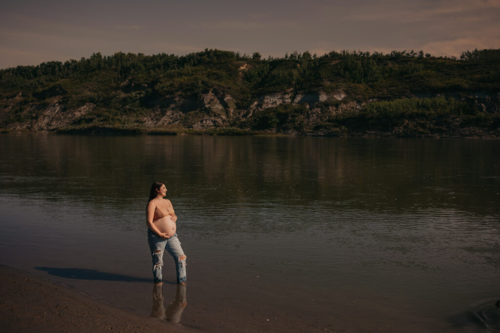 pregnant woman poses in jeans and bra by river during outdoor maternity photos in Edmonton