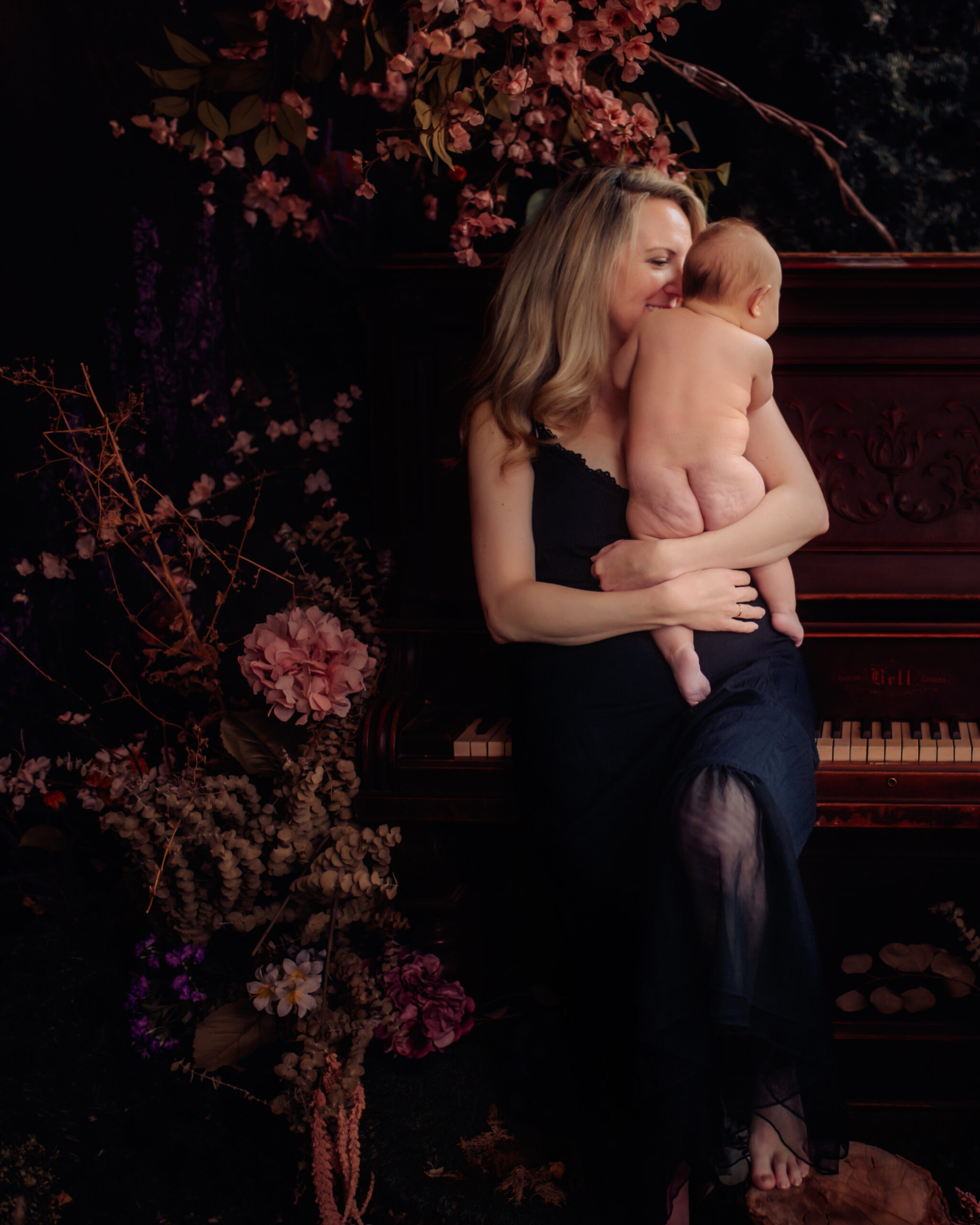 Mother gently holding her baby in a tender embrace, set against a rich floral and piano background, capturing an intimate motherhood moment.