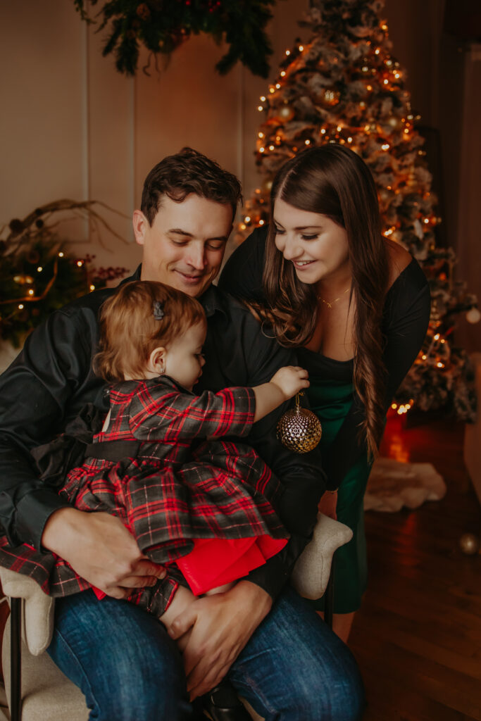 A family sitting together in front of a Christmas tree, with a father holding a young child dressed in a red and black plaid dress and a mother leaning in, smiling warmly as the child holds a gold ornament